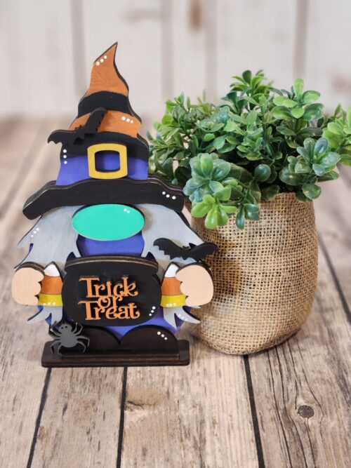 Witchy Standing Gnome, witch hat, candy corn and witches cauldron with Trick or Treat in orange letters, background is a wood table, green plant wrapped in burlap and a wood slat wall.