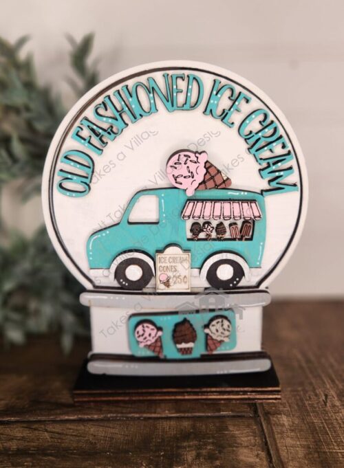 White background with blue "Old Fashioned Ice Cream", blue and pink ice-cream truck insert. Blue sign with three ice cream cones on it. All inside a white and grey snow globe frame on a brown wood table with a green wreath next to it and white Shiplap wall behind.