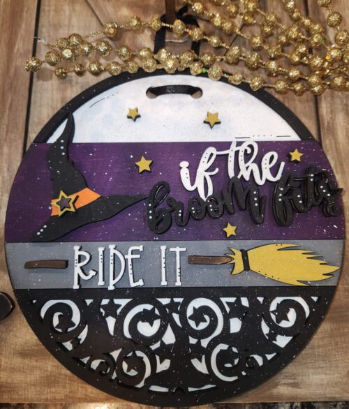 If The Broom Fits Ride It door hanger, black, white and purple, witch hat, broom stick, bottom third witch hat, curlicues and stars cutout pattern. Background is a wood door with gold string of beads.