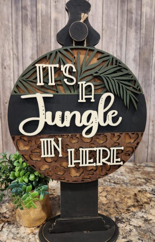 Hanging on a black stand with a plant behind it, standing on a counter with a wood slat wall behind, It's a Jungle In Here door hanger with palm leaf on top wording in center animal print on bottom, interchangeable animal print bottom third, Leopard print, zebra print, giraffe print, DIY Craft kit, or finished product.