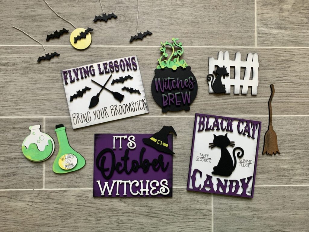 Witches Tier Tray DIY craft set, bats on a string, flying lessons bring your broomstick sign, bubbling cauldron with "Witches Brew" on it, black cat in front of white picket fence, brown broomstick, two beakers with green poison in them, It's October Witches sign, black cat candy sign with a black cat and purple writing, background is a wood look title floor with white grout.