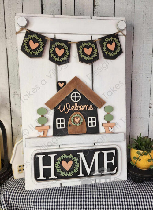 White interchangeable pallet board with home and planter pots insert. Planter pots are tan and bushes are green. home is black with a brown roof and door with a peach "welcome" across the home. Black sign with white "HOME" with a wreath with a pink heart inside replacing the O. Sitting on a black and white gingham fabric with spring décor and white slat wood walls.