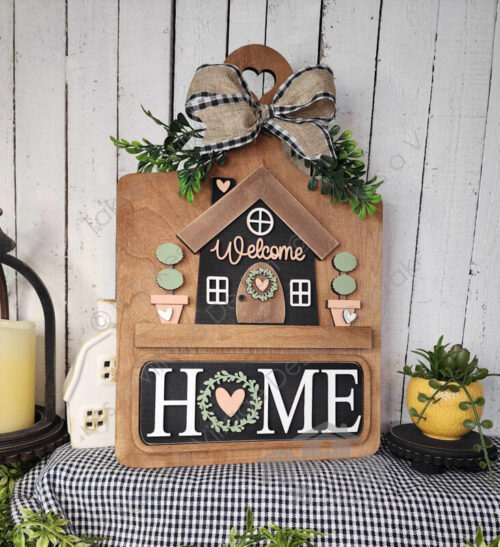 Brown cutting board with home and planter pots insert. Planter pots are tan and bushes are green. home is black with a brown roof and door with a peach "welcome" across the home. Black sign with white "HOME" with a wreath with a pink heart inside replacing the O. Sitting on a black and white gingham fabric with spring décor and white slat wood walls.