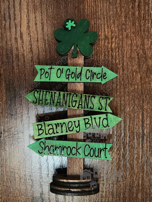 Mini Street Sign Post, Dark Green Shamrock on top of the sign with a small light green shamrock on it. Top direction sign pointing right is green and says Pot O' Gold Circle, Next sign green pointing left Shenanigans St. Third sign green pointing right Blarney BLVD, last pointing left green Shamrock Court.